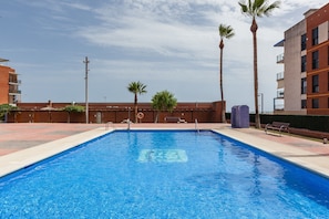 Una piscina rectangular con agua azul cristalina, rodeada por un muro de ladrillo, palmeras y un edificio con balcones.