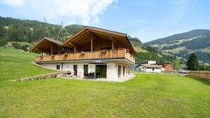 A two-story wooden chalet with a balcony, surrounded by a grassy field and mountains.
