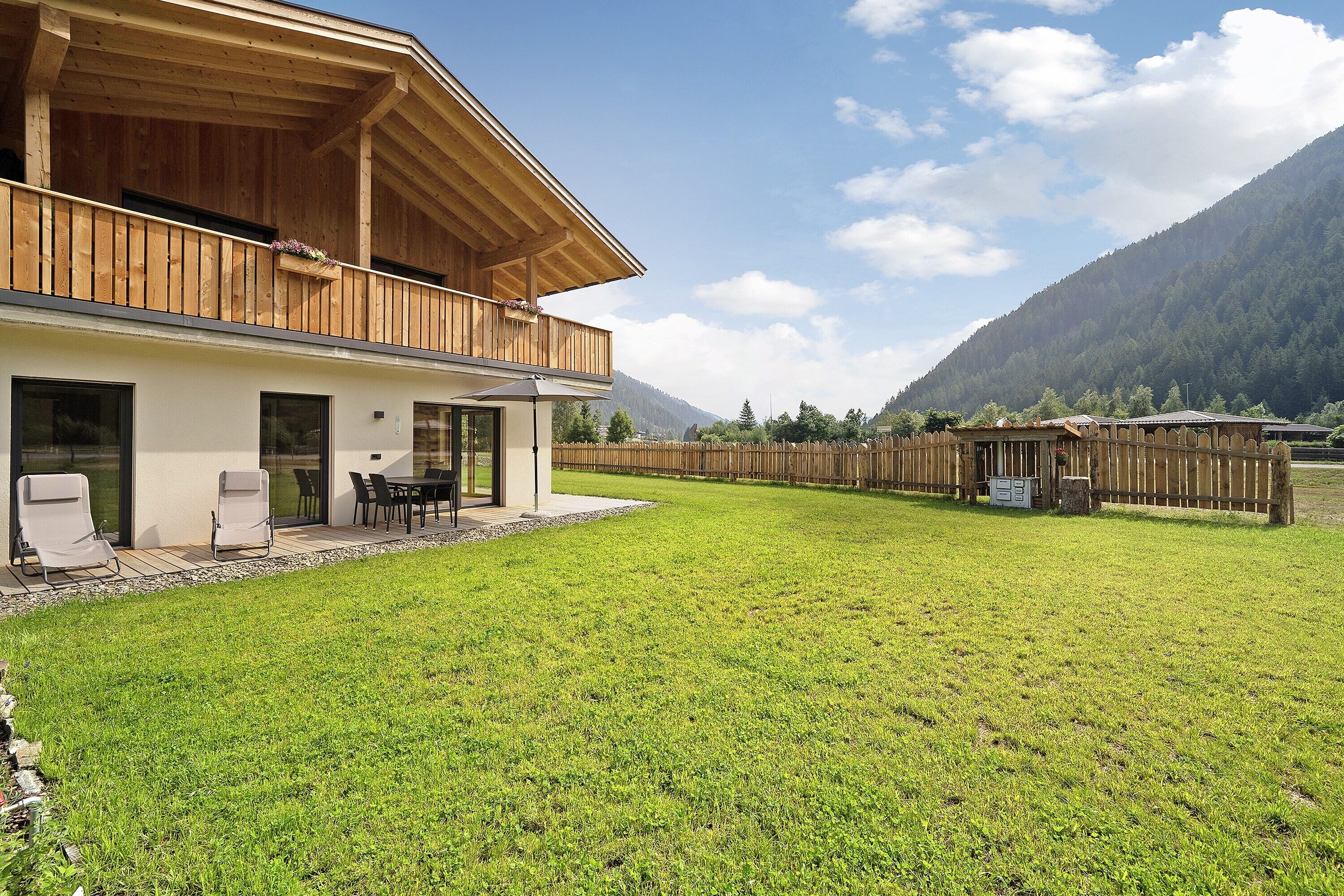 A two-story wooden house with a balcony, a patio with chairs, and a view of a mountain.