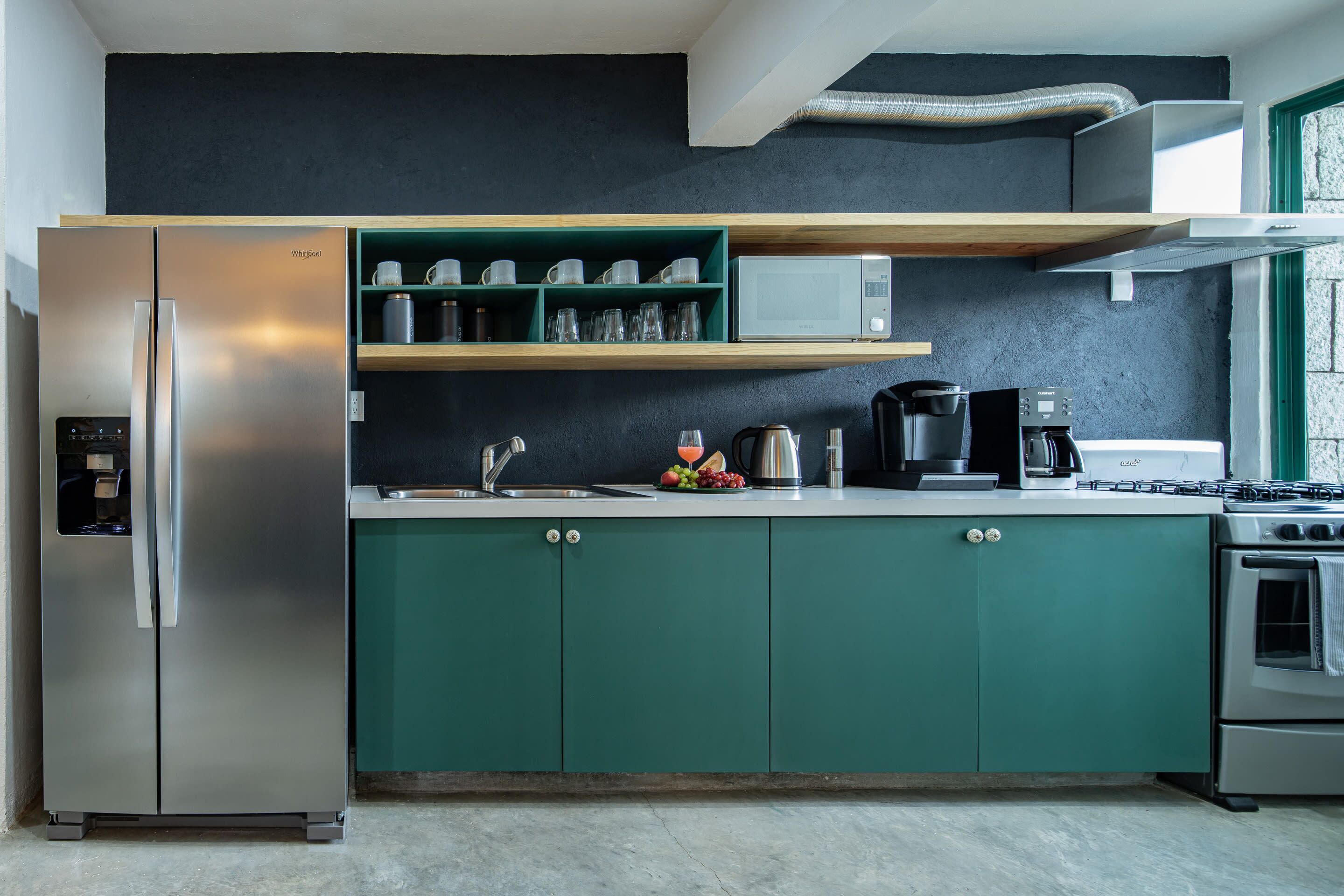 Una cocina moderna con un refrigerador de acero inoxidable, un microondas y una estufa. Hay armarios verdes y una encimera con varios utensilios de cocina.