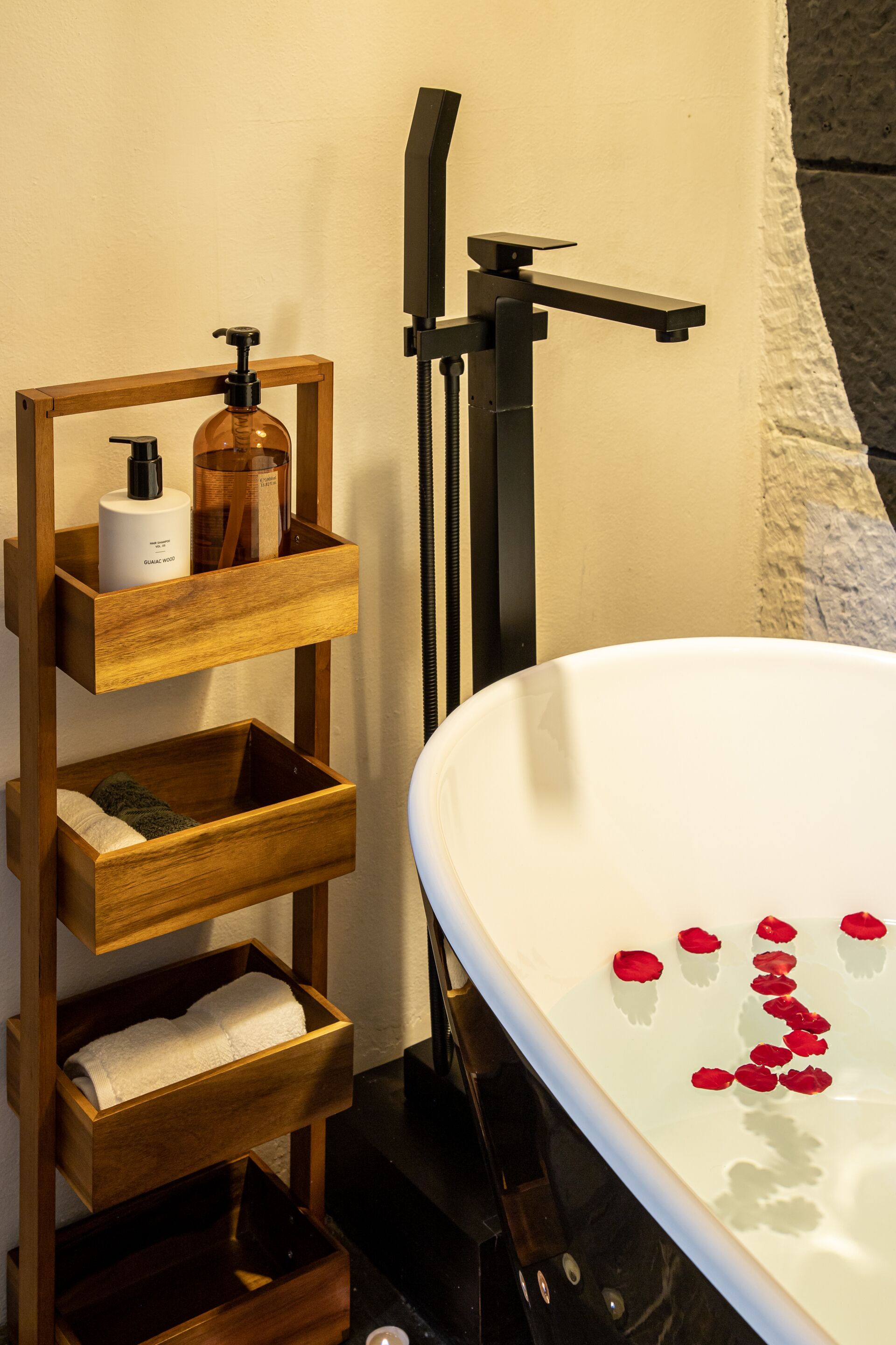 A bathroom with a white bathtub, wooden shelves with toiletries, and rose petals on the tub.