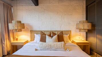 Una cama bien hecha con una colcha blanca y varios almohadones, una mesita de madera con una lámpara pequeña y una vela, y una pared de piedra texturizada detrás de la cama.