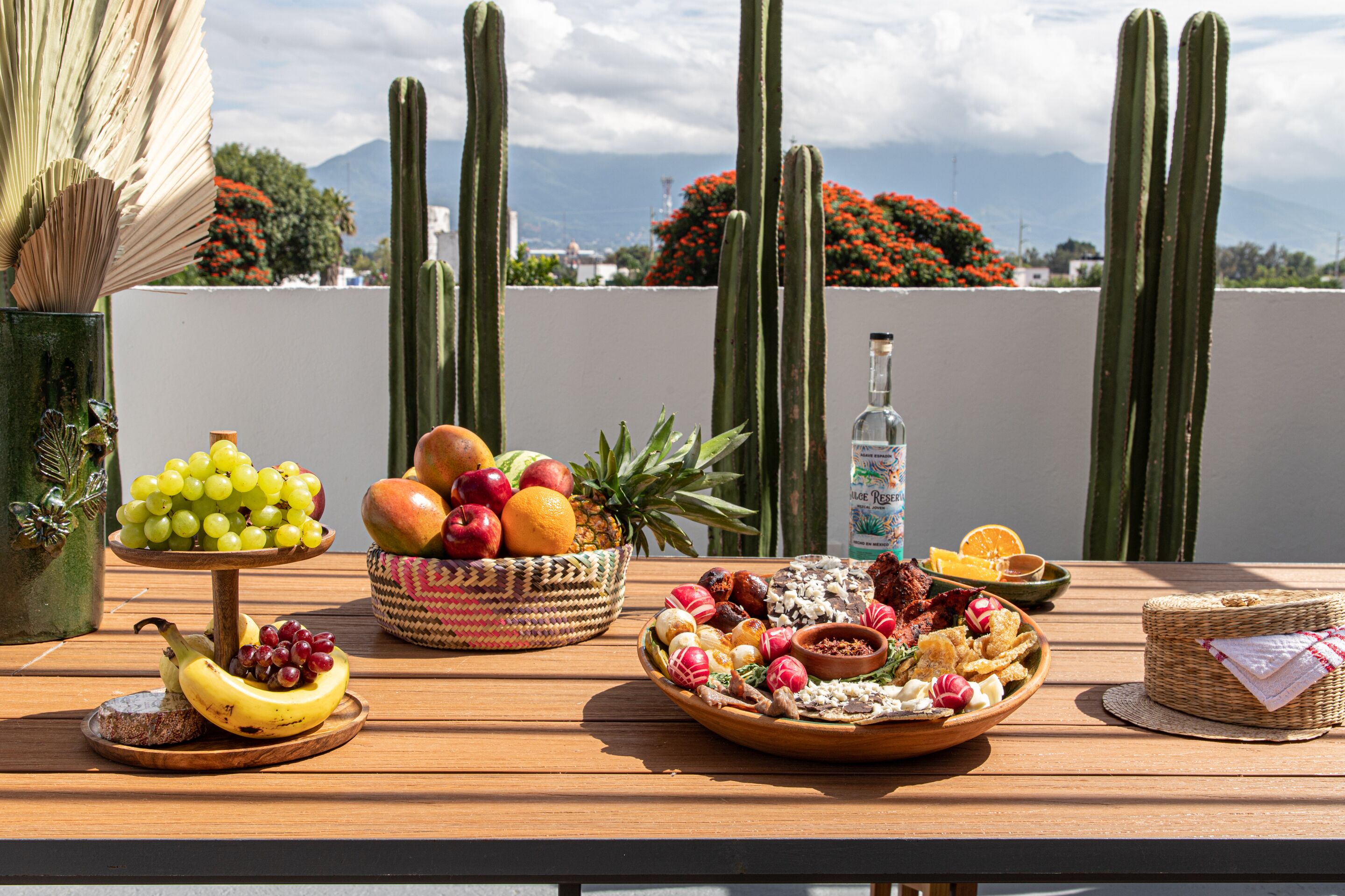 A wooden table with various fruits and snacks, a bottle, and a woven basket on a terrace with cacti and flowers in the background.