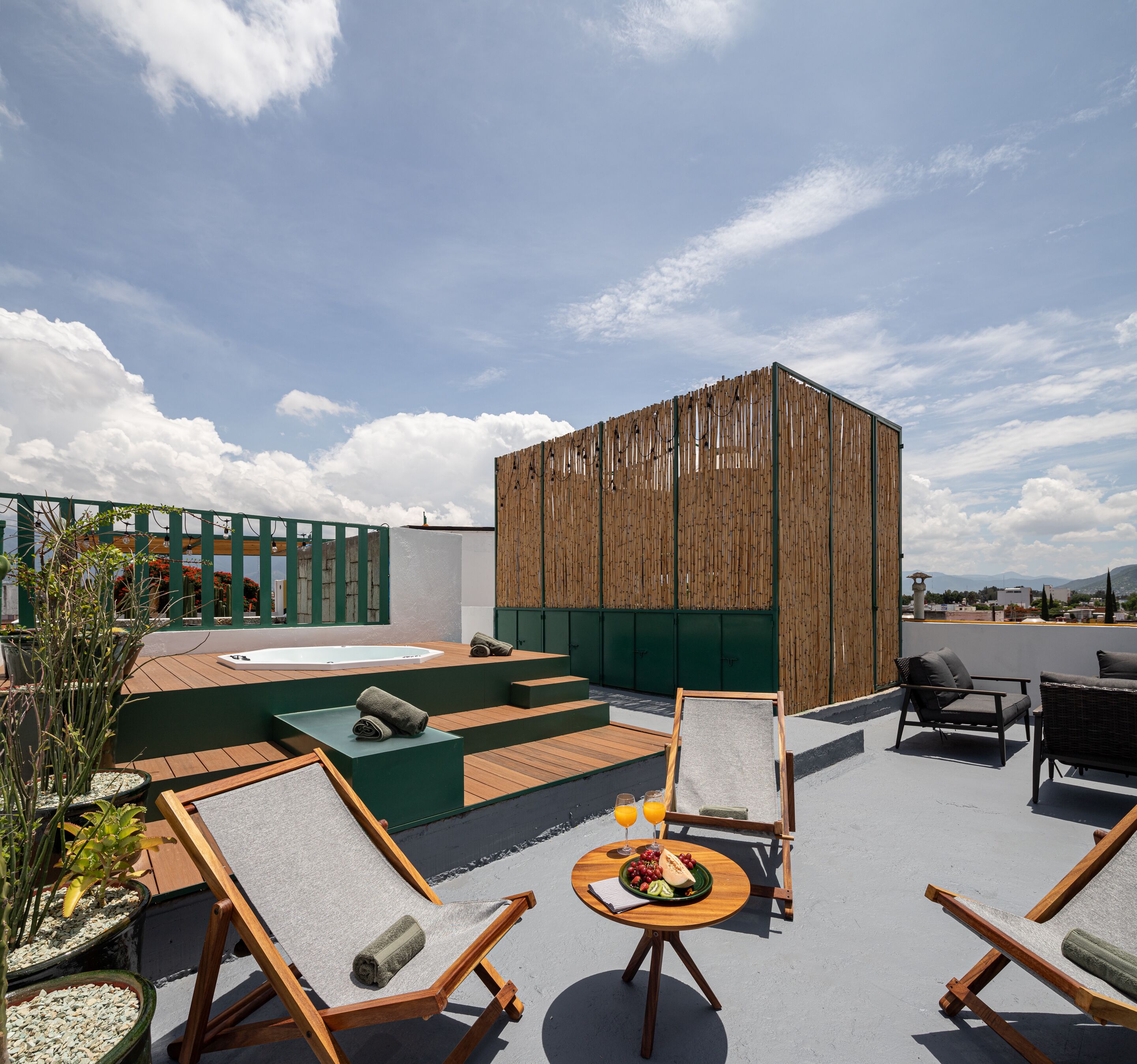 A rooftop terrace with wooden deck, lounge chairs, a table with drinks, and a view of the city.