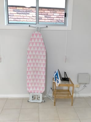 A room with a window, an ironing board, a wooden table with an iron, and a power outlet.