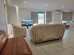 A modern living room with a large flat-screen TV, two covered sofas, a window with a view, and a tiled floor. - Free WIFI, Free Parking, Free over few hundred TV Channel, free Computer Games. (Kingswood)