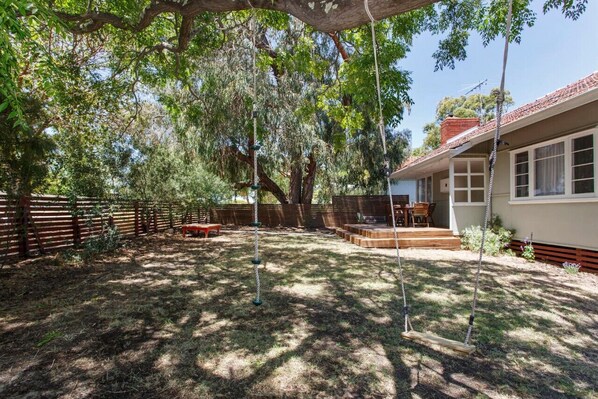 A backyard with a swing set, a wooden deck, and a house with a red roof. - Home Away - 2 Bedroom Cottage (Hilton)