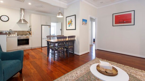 A spacious living area with a wooden floor, a dining table with chairs, a blue sofa, and a kitchen in the background. - Home Away - 2 Bedroom Cottage (Hilton)