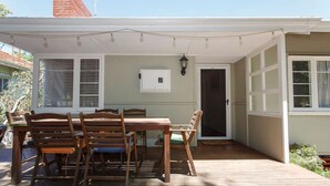 A covered patio with a wooden table and chairs, a white door, and a window with blinds. - Home Away - 2 Bedroom Cottage (Hilton)