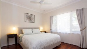 A neatly arranged bedroom with a bed, two bedside tables with lamps, a ceiling fan, a window with curtains, and a framed picture on the wall. - Home Away - 2 Bedroom Cottage (Hilton)