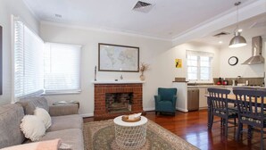 A living room with a fireplace, a dining area with a table and chairs, and a kitchen in the background. - Home Away - 2 Bedroom Cottage (Hilton)
