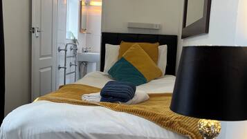 Habitación de hotel con cama, mesita de noche con lámpara, puerta y baño visible a través de una puerta abierta.