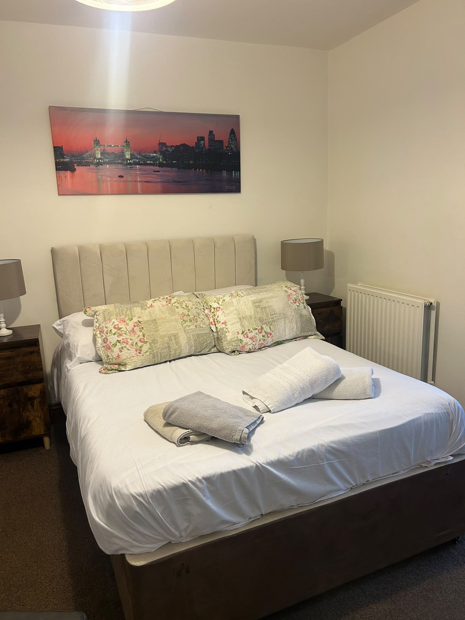 A neatly made bed with floral pillows and a folded towel, a nightstand with a lamp, and a painting of a cityscape on the wall.