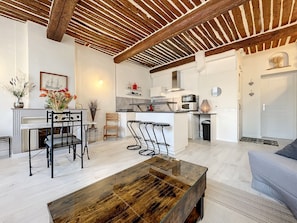 A modern living room with a wooden coffee table, a dining area with chairs, a kitchen with a microwave, and a white door.