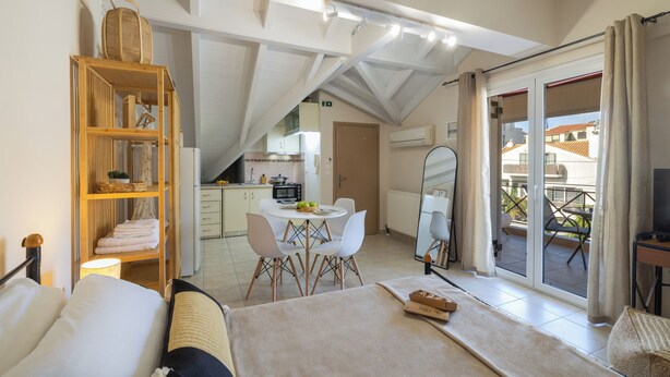 A bedroom with a bed, a bedside table, a wooden shelf with baskets, a dining area with a table and chairs, and a sliding glass door to a balcony. - City Suite - The Loft in Kefalonia (argostoli)