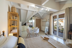 A bedroom with a bed, a bedside table, a wooden shelf with baskets, a dining area with a table and chairs, and a sliding glass door to a balcony.