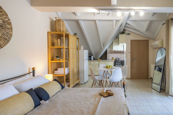 A bedroom with a bed, a wooden wardrobe, a dining area with chairs, a kitchen in the background, and a large mirror.