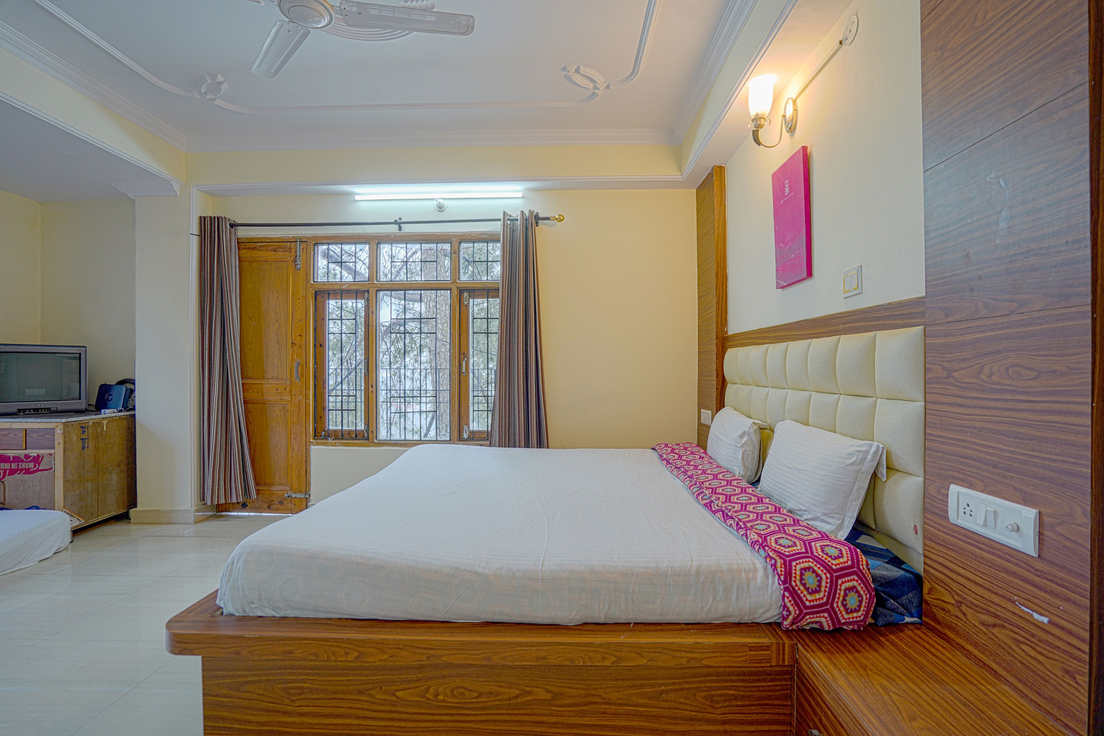 A bedroom with a large bed, wooden headboard, a television on a wooden cabinet, and a window with curtains.