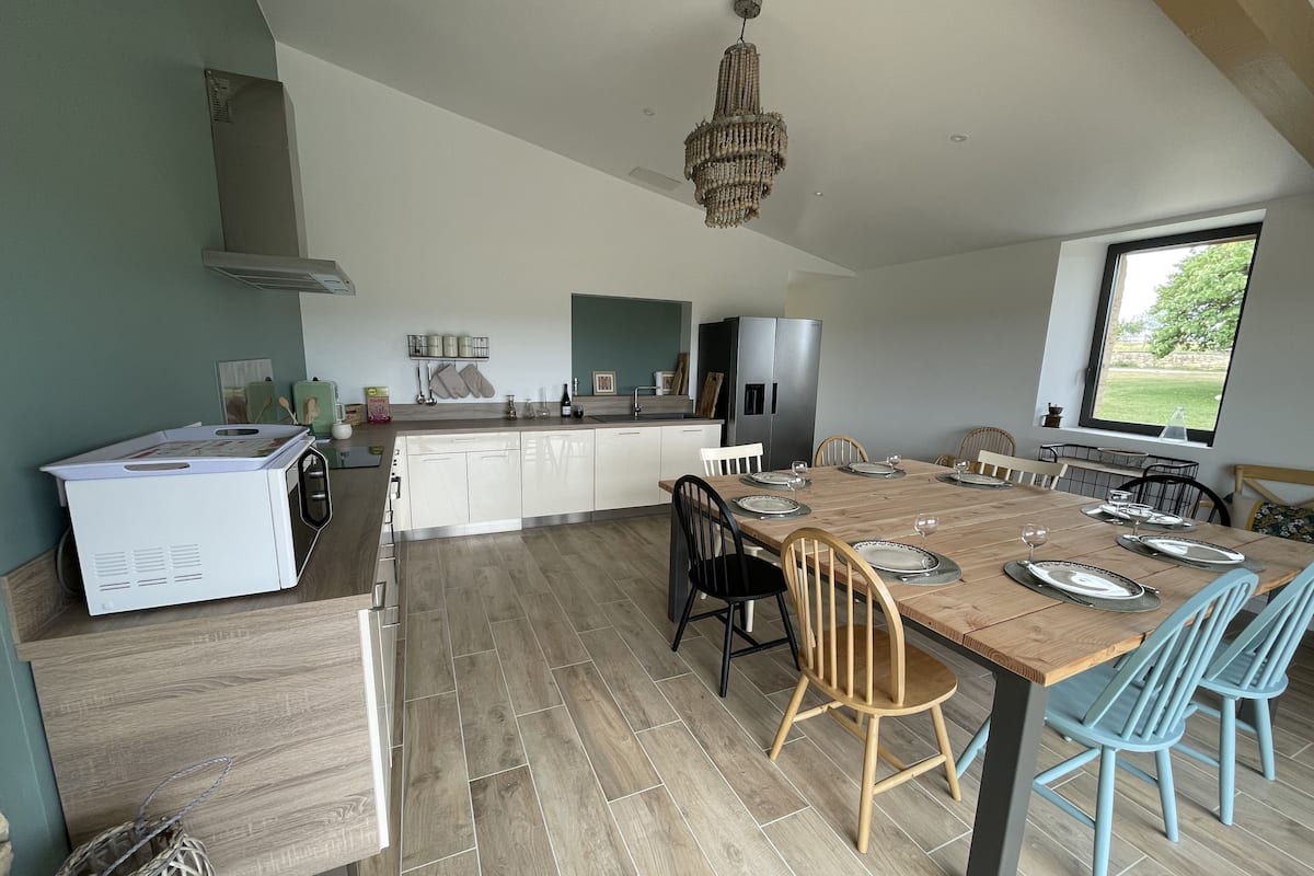 A modern kitchen with white cabinets, a wooden dining table set for six, and a large window providing a view of the outdoors.