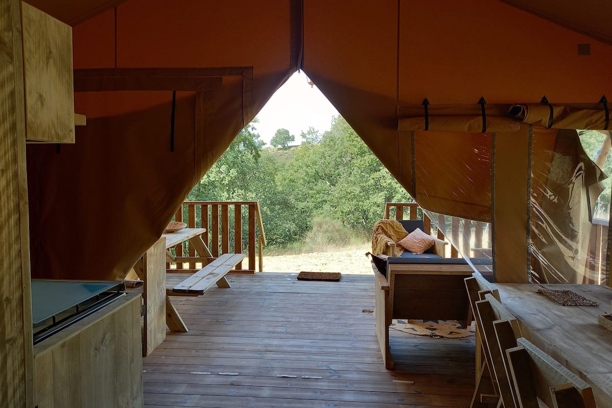 A wooden cabin with a lofted sleeping area, a dining table, and a view of the surrounding forest.