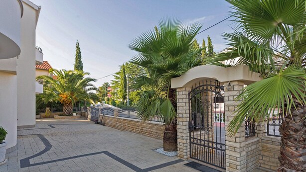 A gated entrance with a stone wall, palm trees, and a paved driveway leading to a building.
