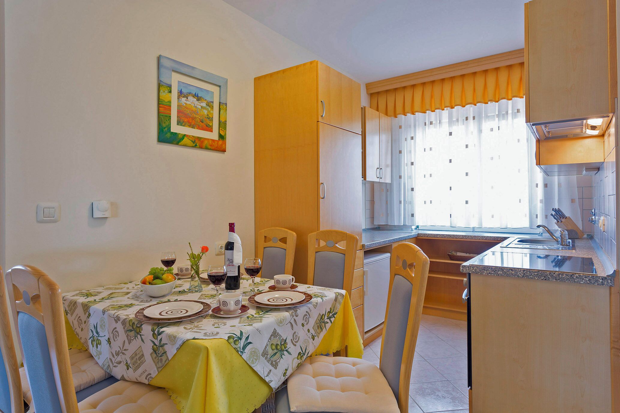 A dining area with a table set for four, a kitchen with wooden cabinets, and a window with curtains.