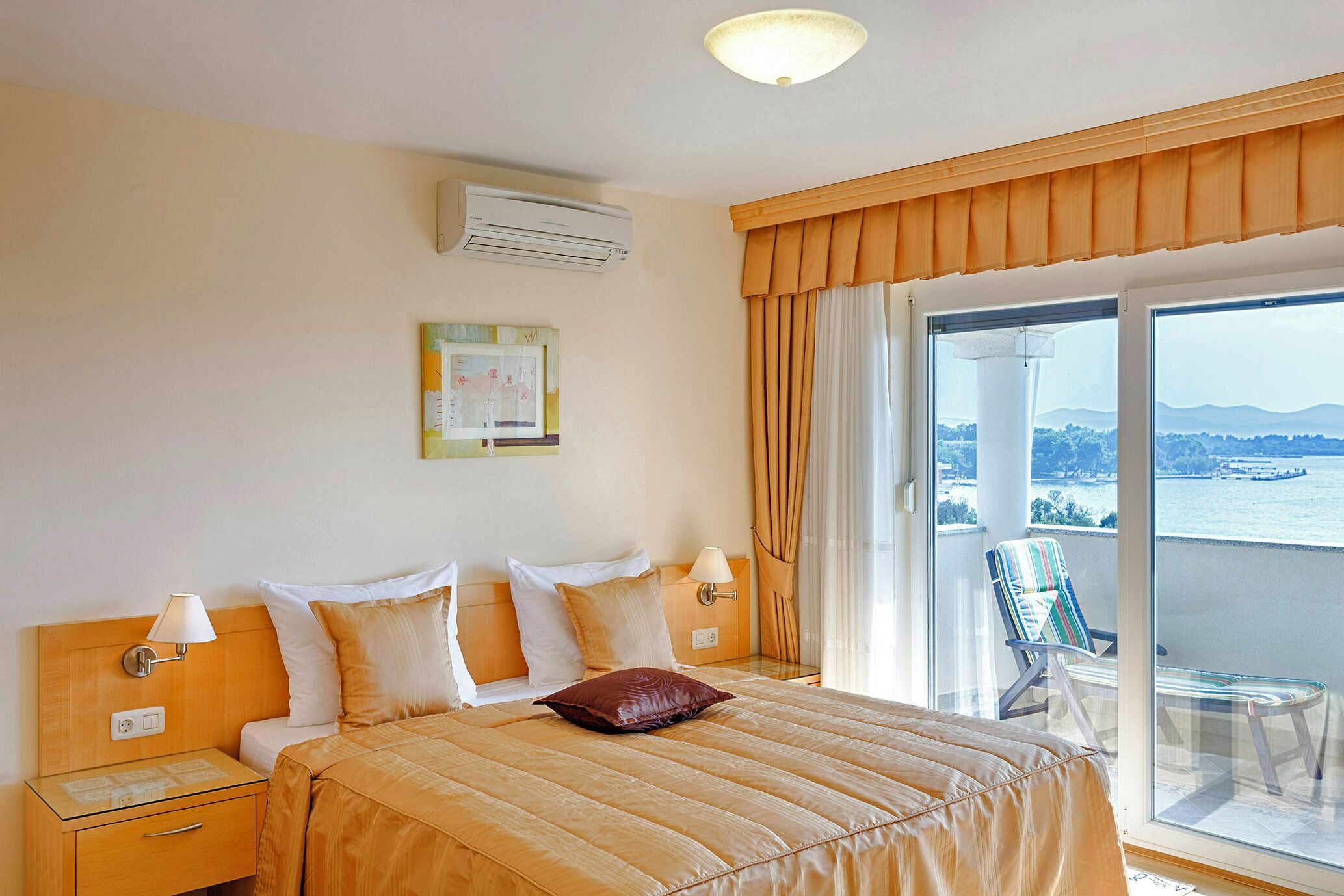 A hotel room with a bed, a nightstand, a lamp, a window with curtains, and a view of a body of water.