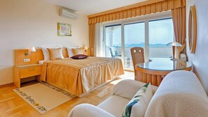 A hotel room with a bed, a small table, a chair, and a view of the sea through large windows.