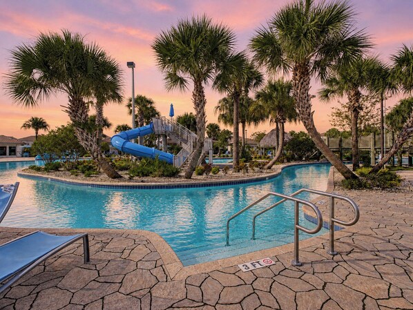 A swimming pool with a blue slide, surrounded by palm trees and a sunset sky.