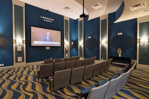 A conference room with a large screen displaying a news broadcast, rows of chairs, and a patterned carpet.