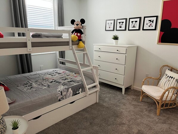 Un lit superposé avec un doudou à l’effigie de Mickey Mouse, une commode blanche, un fauteuil en osier et des photos encadrées accrochées au mur.