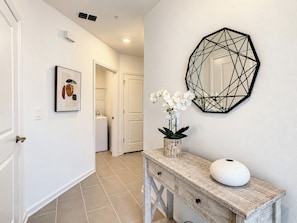 A hallway with a laundry room, a door, a framed picture, a decorative mirror, and a vase with flowers on a wooden console.