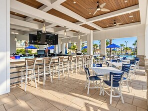 An outdoor patio area with multiple tables and chairs, a bar area with a television, and a pool in the background.