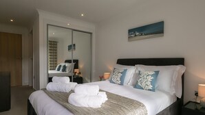 Una cama bien hecha con ropa blanca y almohadones decorativos, una mesita de noche con lámpara, y un espejo grande que refleja la habitación.
