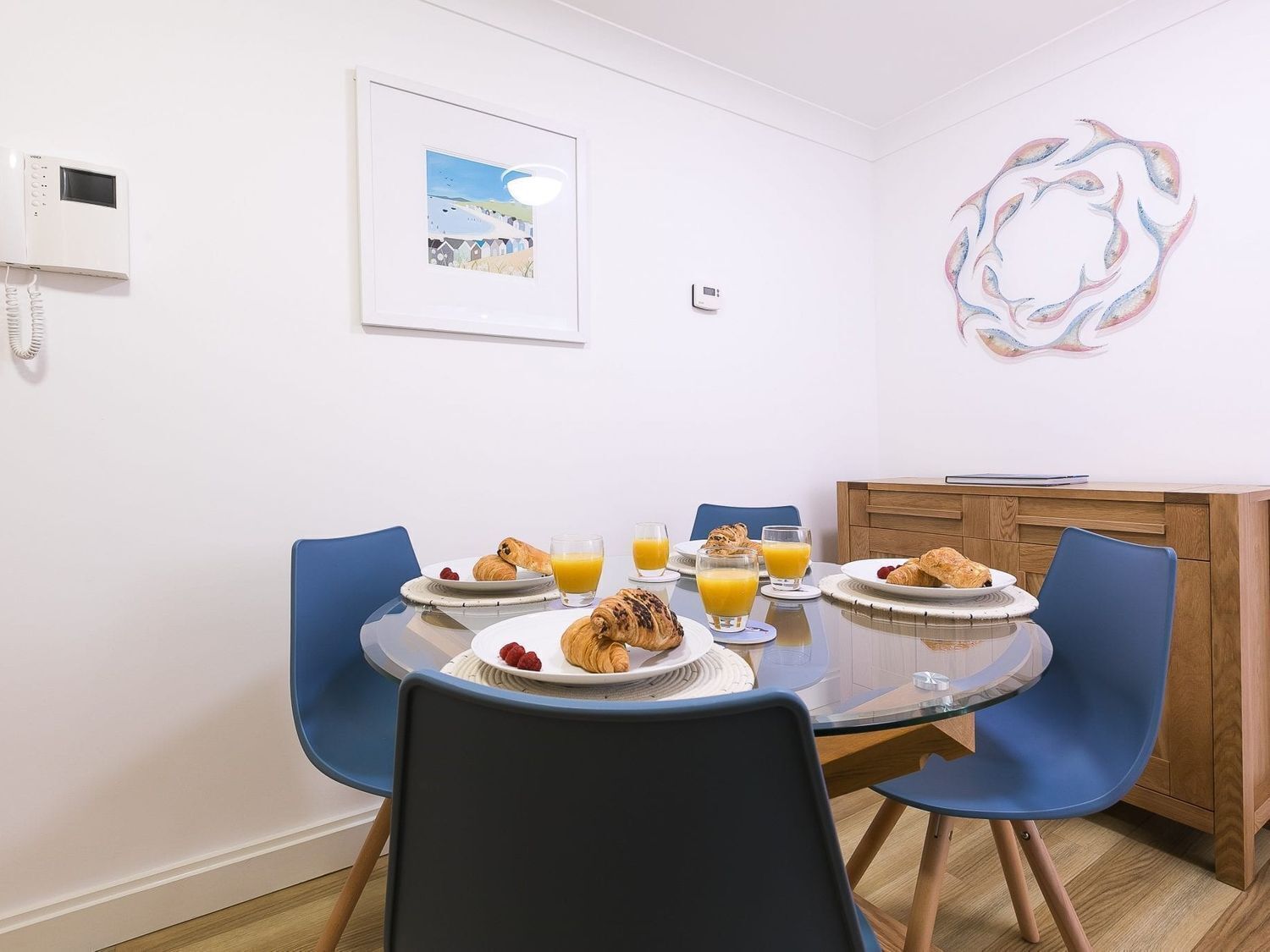 A breakfast setup with croissants, juice, and a plate of raspberries on a glass table with blue chairs.