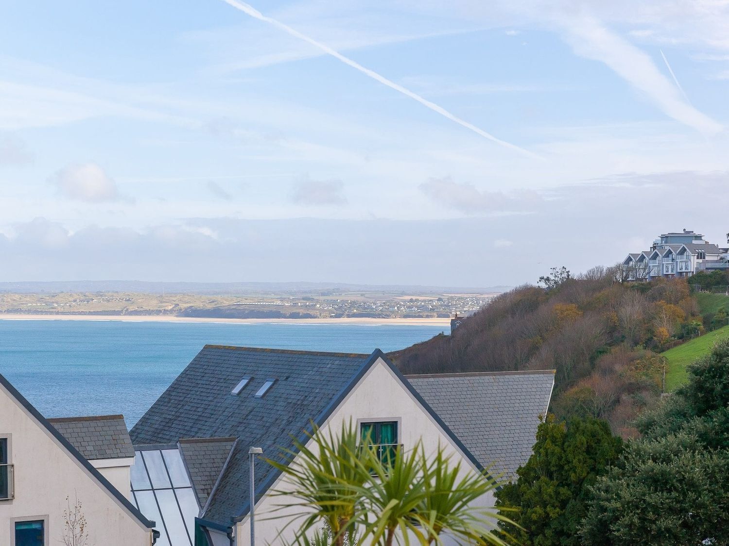 A coastal view with a house overlooking the sea, a beach, and a hill with buildings.