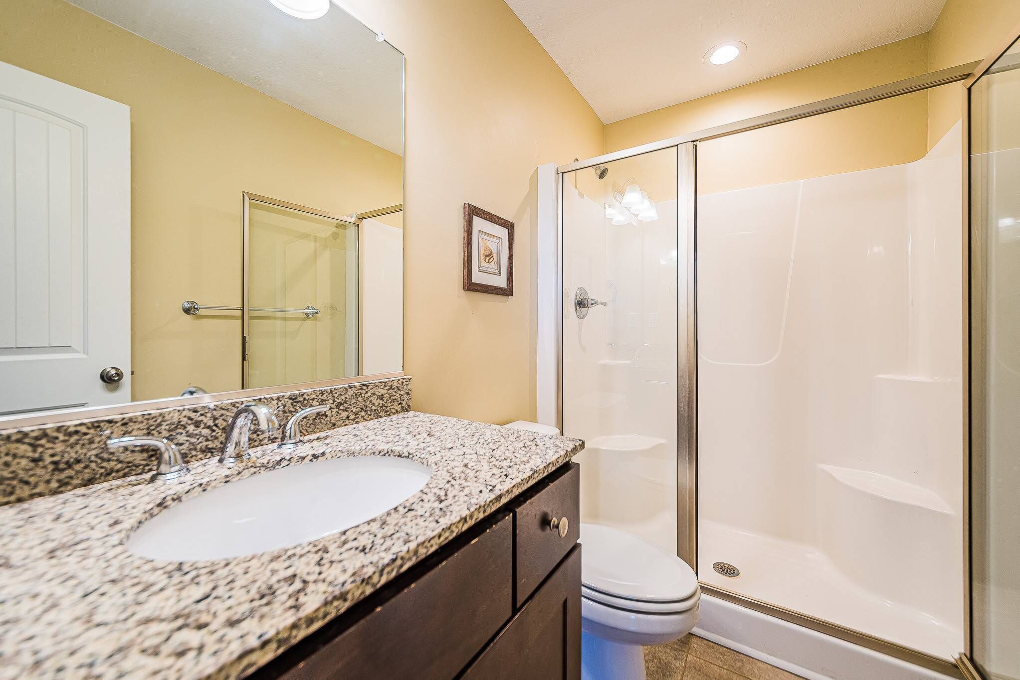 A bathroom with a granite countertop sink, a mirror, a toilet, and a glass-enclosed shower.