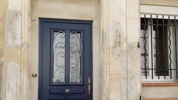 Una puerta azul oscuro con paneles de vidrio decorativos y un tirador de metal, ubicada en un edificio de piedra con una pequeña ventana arriba.