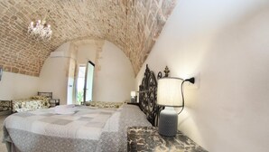 Une chambre avec un plafond en pierre, un lit recouvert d’une couvre-lit à motifs, une table de chevet avec une lampe, et une vue sur l’extérieur par une porte.