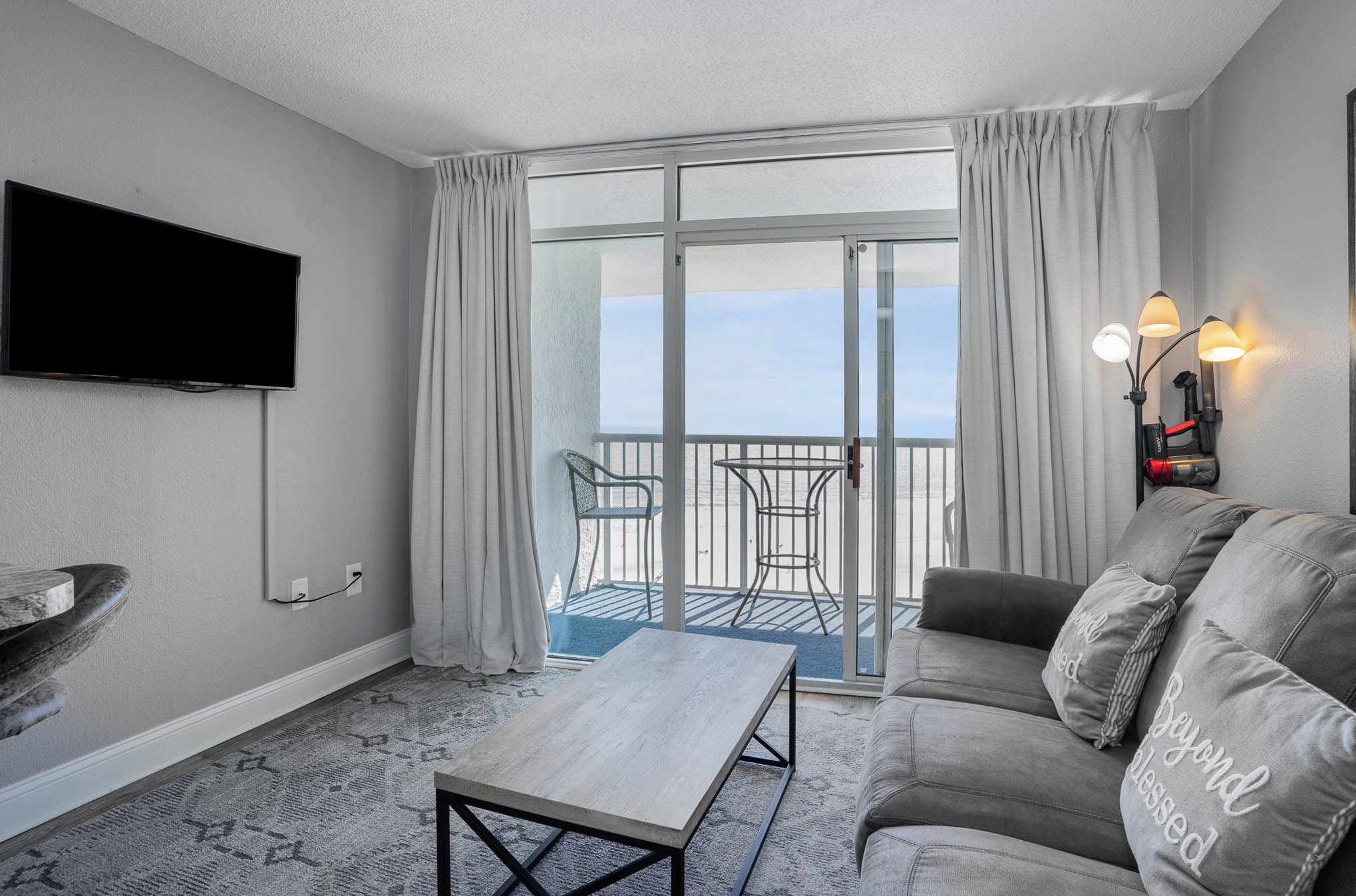 Un soggiorno moderno con una TV a schermo piatto, un divano grigio con cuscini decorativi, un tavolino in legno e un balcone con porta a vetri e tende.