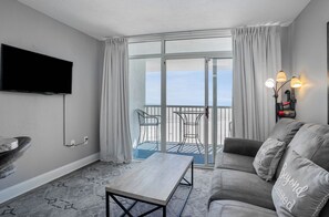 A modern living room with a flat-screen TV, a grey sofa with decorative pillows, a wooden coffee table, and a balcony with a glass door and curtains. - Beautiful Oceanfront 1 Bedroom Condo at Seawatch Resort (Myrtle Beach)