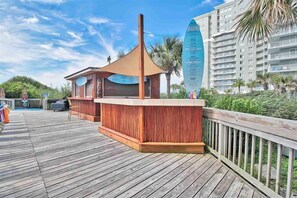 A wooden deck with a bar area, a tall building in the background, and a palm tree. - Beautiful Oceanfront 1 Bedroom Condo at Seawatch Resort (Myrtle Beach)