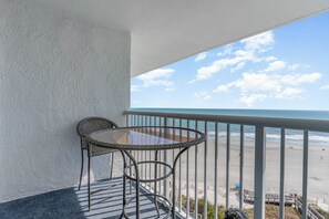 A balcony with a wicker table and chair, overlooking a beach with a clear sky. - Beautiful Oceanfront 1 Bedroom Condo at Seawatch Resort (Myrtle Beach)