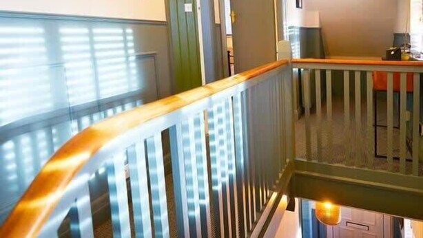 Eine Treppe mit hölzernem Geländer und Metallspindeln, die zu einer Zwischenebene mit einem kleinen Tisch und einem Stuhl führt.