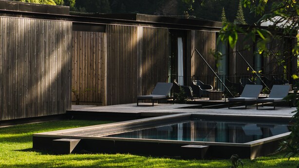 Un área de piscina moderna al aire libre con una terraza de madera, sillones de descanso y vistas a una ladera boscosa.