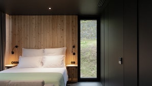 Un dormitorio moderno con cabecera de madera, una cama grande con ropa blanca, un plaid verde, una mesita de noche con lámpara y vistas a un muro de piedra y vegetación al exterior.