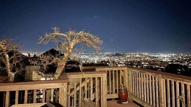 Vue nocturne depuis un balcon donnant sur un paysage urbain, avec une balustrade en bois et un pot de fleurs.