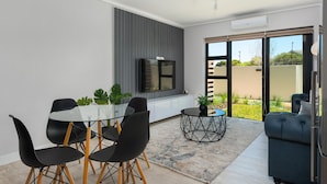 Una sala de estar moderna con una mesa de comedor de vidrio, sillas negras, un sofá y un televisor montado en la pared.