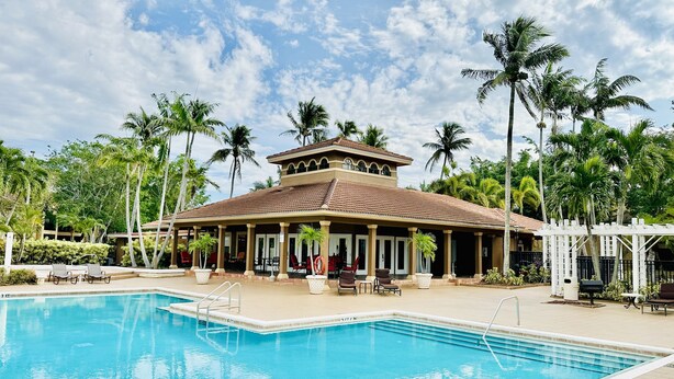 A pool area with a covered seating area, palm trees, and a building with a tiled roof. - Oasis Resort Style Condo 2/2 (Naples)
