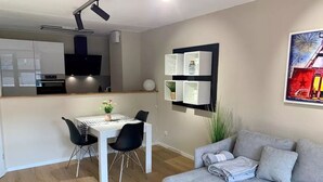 Un salon moderne avec un canapé gris, une table à manger avec des chaises, un coin cuisine et une étagère murale agrémentée d’objets décoratifs.